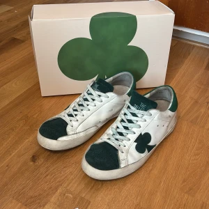 Vita och gröna sneakers från The GALLERIST - Säljer ett par vita sneakers med gröna detaljer och klövertryck på sidan från The GALLERIST. Skorna har snörning, grönt mockaparti vid tån och hälen samt vit platt sula. Perfekta för dig som gillar unik design och streetstyle. SKRIV GÄRNA FÖR BYTEN!