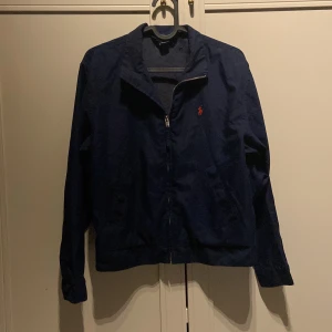 Marinblå vindjacka från Polo Ralph Lauren - Snygg marinblå vindjacka från Polo Ralph Lauren med klassisk röd logga på bröstet. Jackan har dragkedja framtill, två fickor med knapp och en stilren krage. Perfekt för dig som vill ha en clean och tidlös look. 