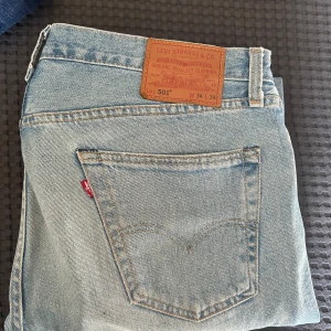 Levi's 501 ljusblå jeans - Klassiska ljusblå Levi's 501 jeans med rak passform och femficksdesign. Jeansen har den ikoniska läderpatchen bak i midjan och röd Levi's-tagg på bakfickan. Perfekta för en avslappnad stil.