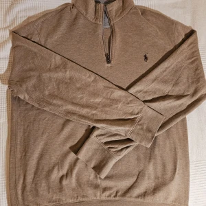 Beige halvzip tröja från Polo Ralph Lauren - Säljer en beige långärmad halvzip tröja från Polo Ralph Lauren med broderad logga på bröstet. Tröjan har ribbade muddar och hög krage med dragkedja. Perfekt för en stilren och avslappnad look.