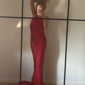 Klänning - Säljer en elegant röd långklänning med halterneck och guld detalj vid halsen.Klänningen har en stilren och enkel design i satin, och är ärmlös med liten öppning i ryggen.