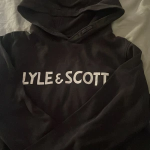 Svart hoodie från Lyle & Scott - Svart hoodie från Lyle & Scott med stor vit logga på bröstet. Klassisk huva och långa ärmar, tillverkad i mjukt material som passar perfekt för en avslappnad stil.