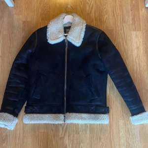 Svart pälsfodrad jacka från ASOS - Säljer en svart jacka från ASOS med vit fuskpäls på krage, ärmslut och nederkant. Jackan har dragkedja framtill, två snedställda fickor och bälte i midjan. Perfekt för kalla dagar och riktigt snygg till jeans.