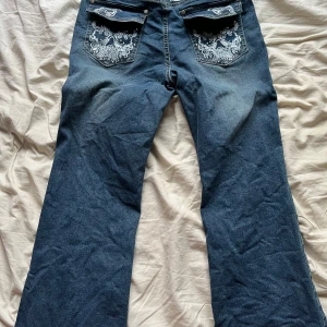 Blå bootcut jeans med broderade bakfickor - Säljer ett par blå bootcut jeans med snygga broderade detaljer på bakfickorna. Jeansen har en klassisk femficksdesign och är perfekta för dig som gillar en lite vidare passform nertill. Aldrig använda 