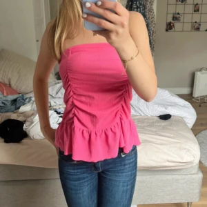 Rosa volang topp från Gina Tricot - Säljer en söt rosa topp från Gina Tricot med volangkant och rynkade detaljer framtill. Toppen är axelbandslös och har en croppad passform som sitter tight. Perfekt för dig som gillar färg och trendiga detaljer!