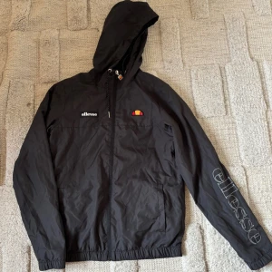 Svart vindjacka från Ellesse - Säljer en svart vindjacka från Ellesse med huva och dragkedja. Jackan har Ellesse-logga på bröstet och ärmen, samt snörning i huvan. Tillverkad i polyester och har meshfoder på insidan. Lite osäker på storleken men tror xs/s