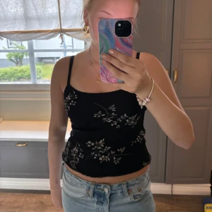 Svart blommig linnetopp från Fashion Maker - Svart linnetopp med tunna axelband och vita blommiga broderier. Toppen är croppad och har vågig kant nertill. Perfekt för varma dagar och enkel att styla med jeans eller kjol.