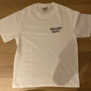 Vit t-shirt från Gallery Dept. - Vit t-shirt från Gallery Dept. med svart tryck på bröstet och stor logga på ryggen. Klassisk rund hals och normal passform. Perfekt för en clean och stilren look.