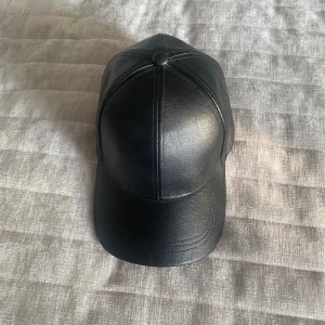 Svart keps i skinnimitation - Snygg svart keps i skinnimitation med klassisk böjd skärm och paneldesign. Kepsen har justerbart spänne baktill för perfekt passform och stilrena sömmar som ger en clean look. Perfekt accessoar för att lyfta din outfit.