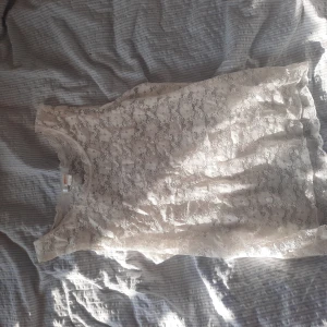 Vit spetslinne med blommönster - Säljer ett vitt ärmlöst linne i spets med blommigt mönster. Perfekt för dig som gillar romantisk och feminin stil. Linnet är lätt och luftigt, med rund halsringning och transparent känsla tack vare spetsmaterialet.