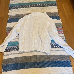 Vit stickad kabeltröja med polokrage - Säljer en vit stickad tröja med klassiskt kabelstickat mönster och hög polokrage. Tröjan har långa ärmar och ribbade muddar, perfekt för kyliga dagar. Enkel att matcha med jeans eller kjol för en clean look.
