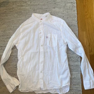 Vit skjorta med ståkrage från Levi's - Säljer en vit skjorta från Levi's med ståkrage och bröstficka. Skjortan har långa ärmar och knäppning framtill. Perfekt för dig som gillar en clean och stilren look.