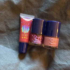 Q-KI läppglans och nagellack set - Säljer ett set med ett rosa läppglans i tub och två nagellack från Q-KI. Ett nagellack är glittrigt rött och det andra är ljusrosa. Alla har blå lock och smidiga förpackningar, perfekta att ta med i väskan.