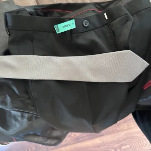 Svart kostym från Dressmann med grå slips - Säljer en svart kostym från Dressmann i slim fit-modell, storlek 52 long och byxa 34-34. Och vit skjorta storlek L .  Kostymen har röda detaljer på insidan och levereras med en ljusgrå slips. Materialmix av polyester, ull och lite lycra för stretch.