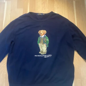 Blå sweatshirt med Polo Bear från Ralph Lauren - Mörkblå sweatshirt från Ralph Lauren med den klassiska Polo Bear-trycket på bröstet. Tröjan har rund halsringning och är långärmad. Perfekt för dig som gillar streetwear och vill ha något bekvämt och stilrent.