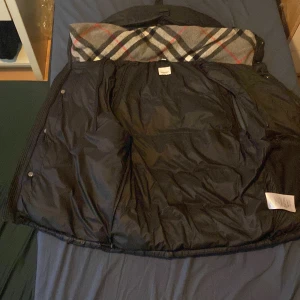 Svart väst / jacka från Burberry - Säljer en svart väst / jacka  från Burberry med avtagbar huva och klassiskt rutigt foder. Västen har knappar och dragkedja framtill samt två fickor med dragkedja. Perfekt för lager-på-lager och snygg detalj med Burberrys rutmönster på insidan.