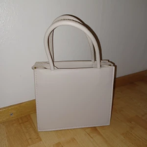 Beige mini handväska med axelrem - Säljer en stilren beige mini handväska med både handtag och lång axelrem. Väskan har en fyrkantig form och är tillverkad i syntetmaterial. Perfekt för dig som vill ha en enkel och trendig accessoar till din outfit.