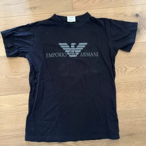 Svart t-shirt med Emporio Armani-tryck - Svart t-shirt med stort Emporio Armani-logga tryckt på bröstet. Klassisk rund hals och korta ärmar. Perfekt för dig som gillar stilrena och ikoniska tryck.