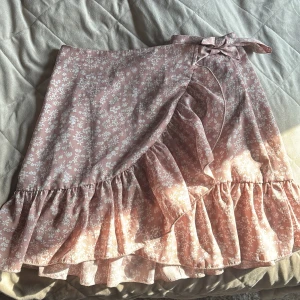 Rosa blommig omlottkjol från SHEIN - Supersöt rosa omlottkjol från SHEIN med vitt blommönster, volangkant och knytband i midjan. Perfekt för dig som gillar romantisk och feminin stil. Kjolen är kort och har en flowy känsla som passar perfekt till sneakers eller sandaler.