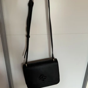 Svart axelväska från Guess - Stilren svart axelväska från Guess i skinnimitation med justerbar rem och diskret logga på framsidan. Väskan har ett ljust innerfoder med Guess-tryck och ett praktiskt innerfack med dragkedja. Perfekt storlek för det viktigaste.