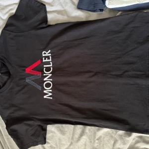 Svart Moncler t-shirt med broderi - Svart t-shirt från Moncler med stort broderat Moncler-logo på bröstet i vitt, rött och blått. Klassisk rund hals och korta ärmar. Tillverkad i mjuk bomull som känns skön mot huden. Perfekt för dig som gillar stilrena och exklusiva plagg.