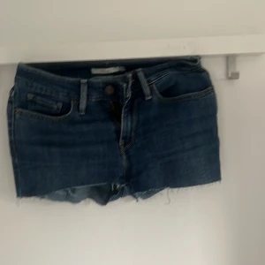 Mörkblå jeansshorts från Levi's - Säljer ett par klassiska mörkblå jeansshorts från Levi's, modell 712 Slim. Shortsen har råa kanter nertill och femficksdesign. Perfekta för sommaren och enkla att matcha med allt.