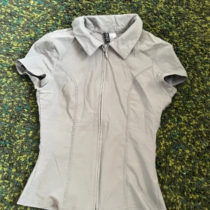Grå kortärmad tight skjorta-grej typ med dragkedja från divided by H&M - Säljer en ljusgrå kortärmad skjorta från divited by H&M med dragkedja framtill och klassisk krage. Skjortan har figurnära passform och är tillverkad i mjuk bomull. Perfekt för en stilren och enkel look.