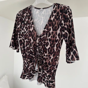 Leopardmönstrad topp med knyt - Säljer en leopardmönstrad topp med djup v-ringning och knyt framtill. Toppen har trekvartsärm och är figurnära i modellen. Perfekt för dig som vill sticka ut med ett trendigt mönster.