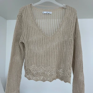 Guldig glittrig stickad v-ringad tröja från NA-KD - Säljer en glittrig guldig/beige stickad tröja från NA-KD med v-ringning och lång ärm. Tröjan har ett genombrutet mönster och dekorativ kant nertill. Perfekt för lager-på-lager och ger en chill vibe.