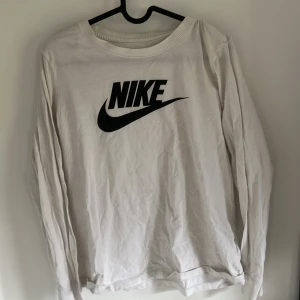 Vit långärmad tröja från Nike - Säljer en vit långärmad tröja från Nike med klassisk svart logga på bröstet. Tröjan är i bomull och har en avslappnad passform, perfekt för en sportig och stilren look.