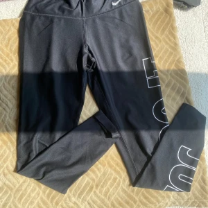 Svarta Nike leggings med tryck - Svarta leggings från Nike med vit text 'JUST DO IT.' längs ena benet. De har en tight passform och är tillverkade i ett stretchigt syntetmaterial, perfekta för träning eller sportiga outfits.