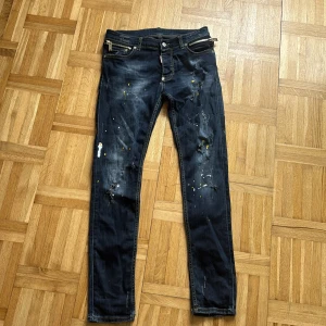 Mörkblå Dsquared2 jeans med dragkedjor - Snygga mörkblå jeans från Dsquared2 med slitna detaljer, färgstänk i gult och vitt samt coola dragkedjor vid fickorna. Jeansen har smal passform och klassisk femficksdesign. Perfekta för dig som gillar streetstyle och unika plagg.