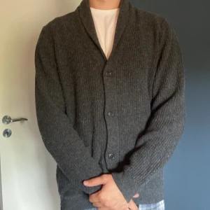 Mörkgrå kofta från Dressmann - Mysig mörkgrå kofta från Dressmann i storlek L. Koftan är ribbstickad med knappar framtill och har en klassisk krage. Perfekt för lager-på-lager och chill höst- eller vinterstil. Passar dig som gillar en enkel och stilren look.