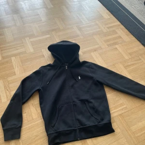 Polo Ralph Lauren zip hoodie svart - Säljer en svart hoodie från Polo Ralph Lauren med dragkedja och huva. Mycket bra skick, använt fåtal gånger. Pyttelite slitage längst upp på dragkedjan, knappt märkbart vid användning - därav det billiga priset. 