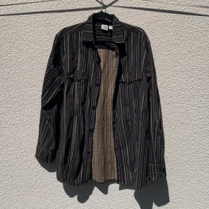 Randig skjorta från 725 Originals - Säljer en långärmad skjorta från 725 Originals i storlek L. Skjortan är randig i svart, beige och blå toner, har knappar framtill och två bröstfickor med knapp. Tillverkad i 100% bomull, och lätt oversized i passformen.