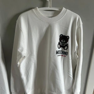 Vit sweatshirt med björntryck från Moschino - Säljer en vit sweatshirt från Moschino med ett stort tryck av en brun björn och texten 'TOY' på bröstet. Tröjan har rund halsringning och långa ärmar. Perfekt för dig som gillar lekfulla designer och streetwear.