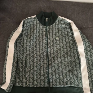 Grön mönstrad track jacket från Lacoste - Säljer en grön track jacket från Lacoste med vitt geometriskt mönster och vita ränder längs ärmarna. Jackan har dragkedja framtill, ribbad krage och muddar samt den klassiska Lacoste-loggan på bröstet.