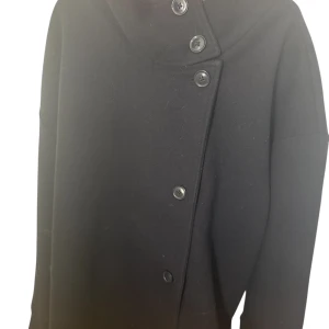 Svart trench coat - Stilren svart kappa i ull med hög krage och snygg asymmetrisk knäppning framtill köpt från Gina tricot. Jackan har långa ärmar och en minimalistisk design som ger en clean look. Perfekt för dig som gillar klassiska vibes med en twist. Den är väldigt stor på mig så säljer den nu. 