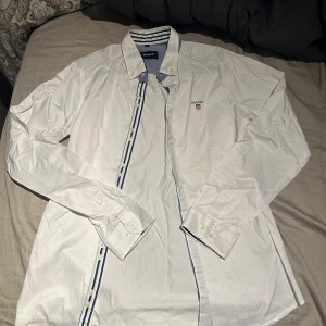 Vit skjorta från Gant med blå detaljer - Vit skjorta från Gant med blå och vita ränder på insidan av kragen och längs knappslån. Klassisk button-down krage och broderad Gant-logga på bröstet. Långärmad och tillverkad i mjuk bomull. Perfekt för en clean och stilren look.