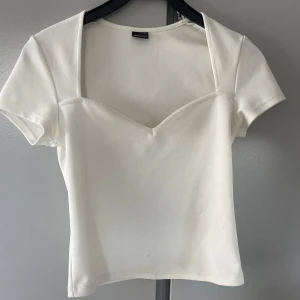 Vit topp med halsringning - Säljer en stilren vit topp med korta ärmar och en snygg v-ringad halsringning. Toppen har en figurnära passform och är tillverkad i ett mjukt material som känns skönt mot huden. Perfekt att matcha med jeans eller kjol. Den har dock en liten fläck längst ner på framsidan, men den går bort vid tvätt💖💖