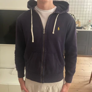 Marinblå hoodie från Polo Ralph Lauren - Säljer en marinblå hoodie från Polo Ralph Lauren med dragkedja och huva. Tröjan har vita snören och den klassiska gula broderade loggan på bröstet. Perfekt för en avslappnad stil.