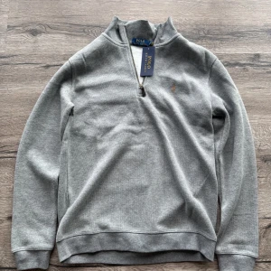 Half zip tröja Polo Ralph Lauren - Snygg grå långärmad tröja från Polo Ralph Lauren med half zip och klassisk broderad logga på bröstet. Tröjan har en hög krage och är gjord i mjuk bomullsmix, perfekt för lager-på-lager. Enkel och stilren design som funkar till allt. Helt ny aldrig använd.