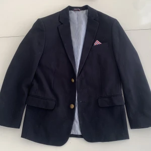 Mörkblå kavaj från Tommy Hilfiger - Stilren mörkblå kavaj från Tommy Hilfiger med klassisk passform. Kavajen har guldfärgade knappar, fickor med lock och ett randigt innerfoder i blått och vitt. Snygg detalj med rutig bröstnäsduk i rött, vitt och blått. Perfekt för en dressad look.