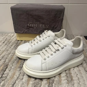 Vita sneakers från Alexander McQueen - Säljer ett par klassiska vita sneakers från Alexander McQueen med tjock sula och diskret guldfärgad logga på hälen. Skorna har rund tå och snörning, perfekta för en stilren look.