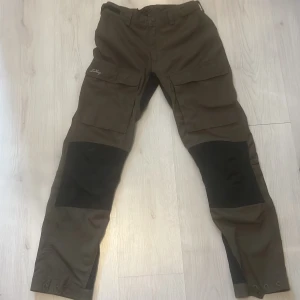 Bruna cargopants från Lundhags - Säljer ett par bruna cargopants från Lundhags med svarta förstärkningar på knäna. Byxorna har stora fickor på benen och är perfekta för uteliv. Tillverkade i slitstarkt material och har justerbara benslut.
