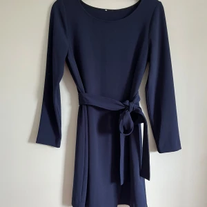 Marinblå klänning med bälte från Twist & Tango - Snygg marinblå klänning från Twist & Tango med lång ärm och bälte i midjan. Klänningen har en rund halsringning och är något stretchig i materialet. Perfekt för dig som gillar enkel och stilren design.