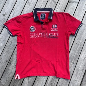 Röd pikétröja från Vinson Polo Club - Röd pikétröja med broderade detaljer och tryck från Vinson Polo Club. Tröjan har klassisk krage i mörkblått med vita ränder, korta ärmar och nummer 26 på ryggen. Perfekt för en sportig och stilren look.