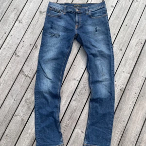 Blå Nudie Jeans - Säljer ett par riktigt snygga klassiska blå jeans från Nudie Jeans med raka ben och snygga slitningar framtill. Jeansen har orange sömmar, fem fickor och är tillverkade i ekologisk denim. Perfekta för en avslappnad stil.