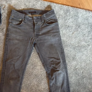 Grå jeans från Nudie Jeans - Säljer ett par grå jeans från Nudie Jeans med klassisk femficksdesign och svarta detaljer på bakfickorna. Jeansen har normal passform och raka ben, perfekta till vardags. Midjan är midwaist och materialet är denim.