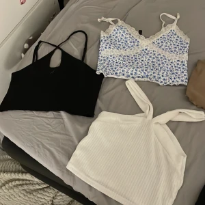 Mix av crop tops i olika färger - Säljer ett paket med sex olika korta crop tops: två svarta, en vit ribbad med twistade axelband, en beige, samt två vita med blått blommönster och spetsdetaljer. Alla är ärmlösa och perfekta för sommaren. Snygga att matcha med jeans eller kjol.